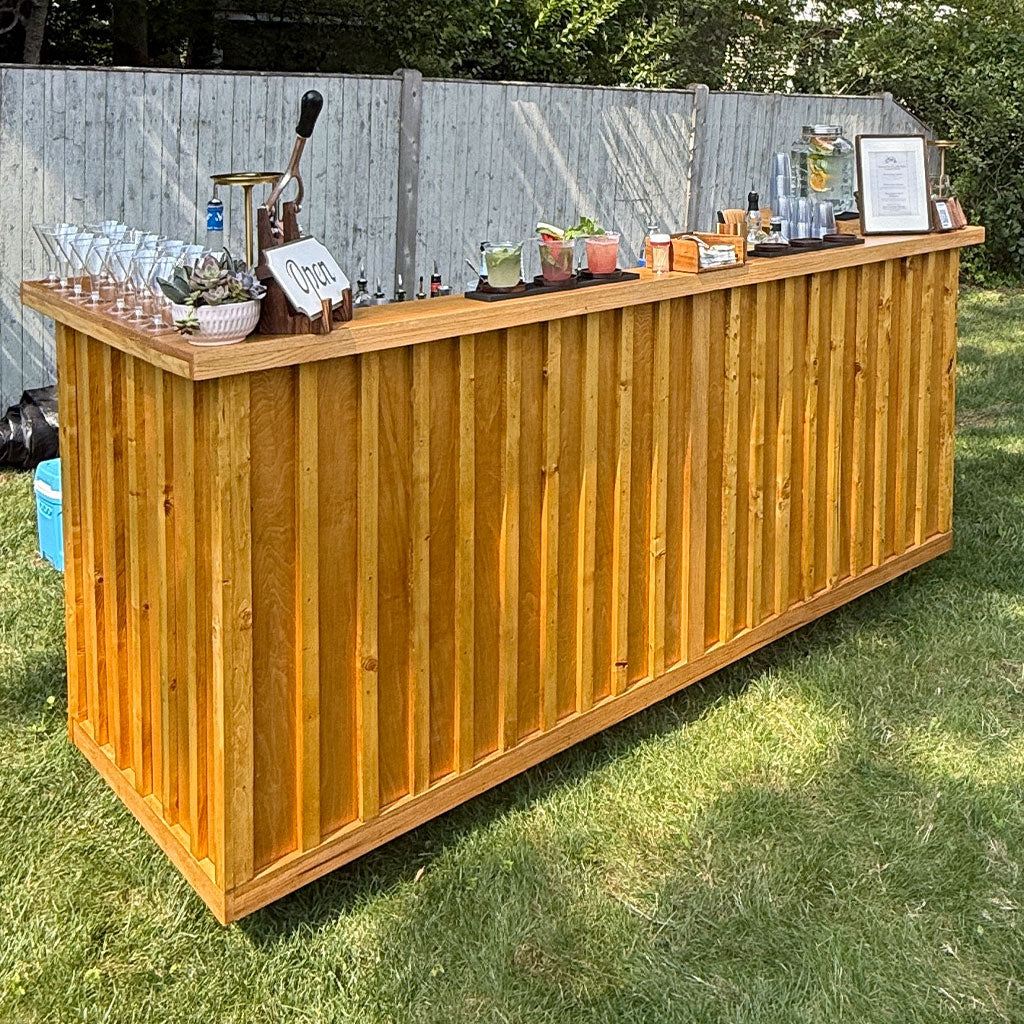 Mobile bar Massachusetts featuring A long wooden outdoor bar counter with a slatted front, set on a grassy lawn in front of a wooden fence. The counter is styled with accessories including glasses, a tap handle, small plants, and signs.