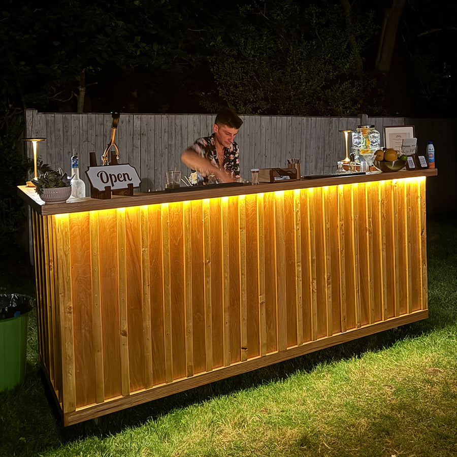 BZ red oak mobile bar lit with warm LED underlighting at a nighttime backyard event, mobile bar rental and dry hire bartending in Duxbury MA